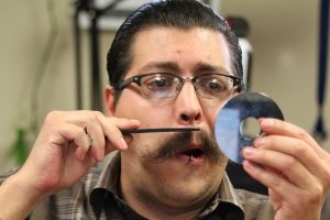 man with glasses and moustashe trimming it with scissors using a cd as a mirror